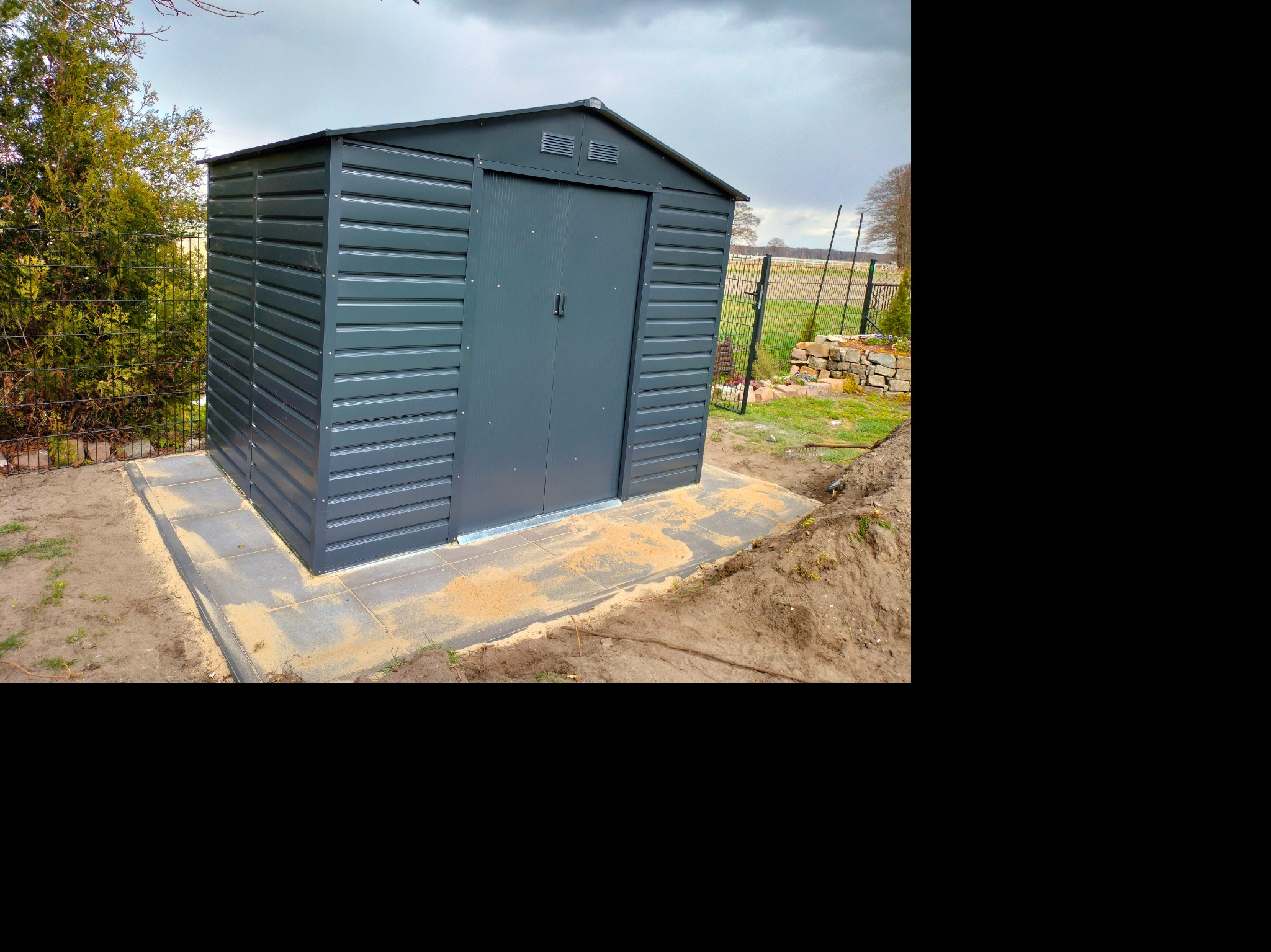 Szara, metalowa szopa ogrodowa z dwuspadowym dachem, umieszczona na betonowej podstawie z kostki brukowej, otoczona trawnikiem i ziemią, widoczna siatka ogrodzeniowa w tle.