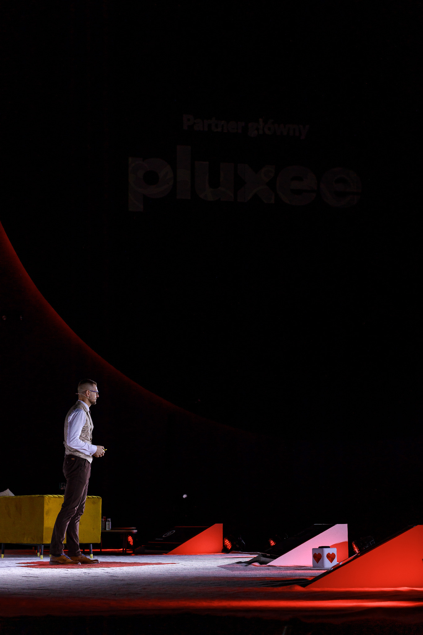 Mężczyzna w kamizelce i okularach stoi na scenie podczas prezentacji, w tle logo 'pluxee' i elementy scenografii.