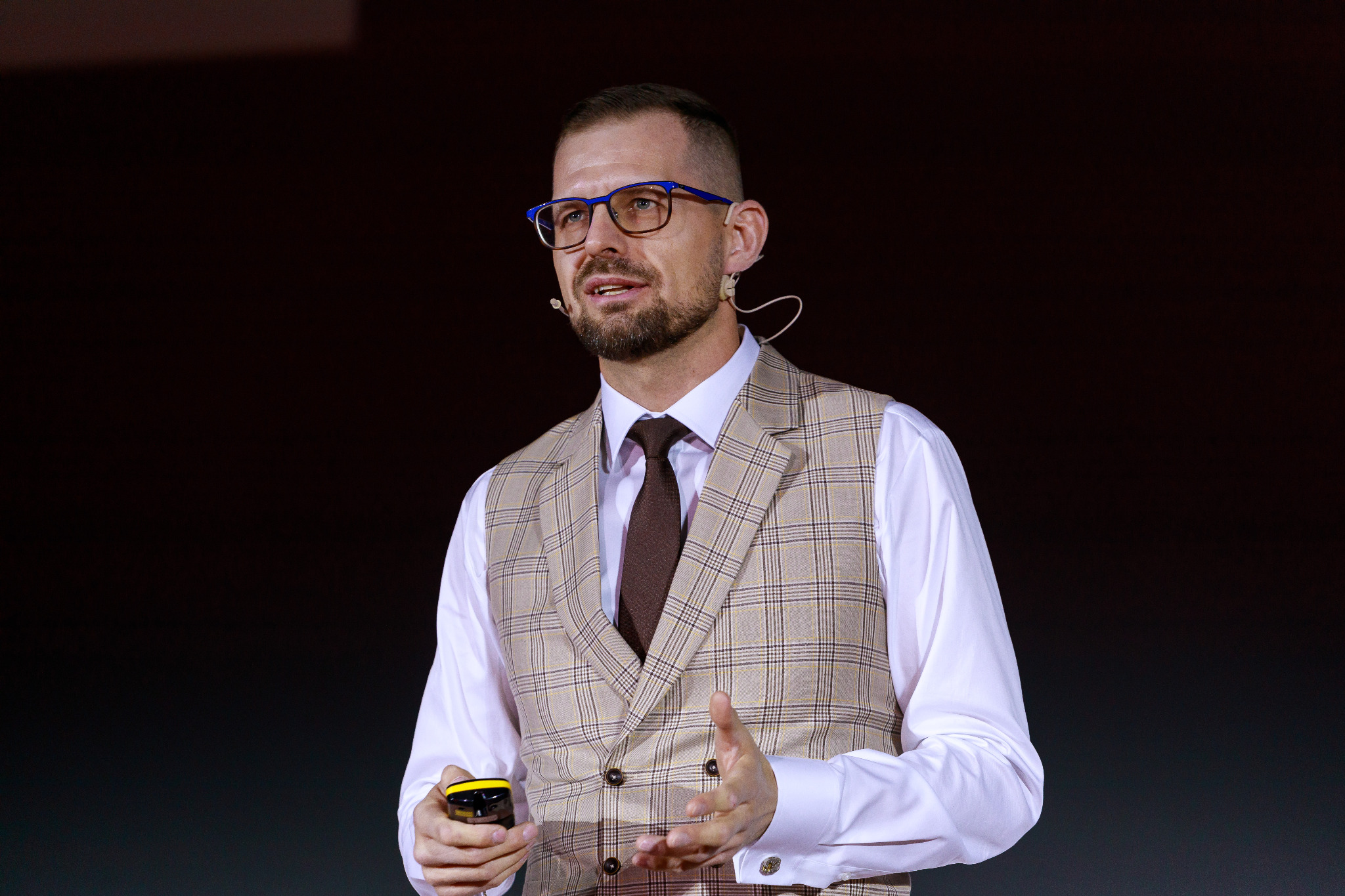 Prelegent w okularach i kamizelce w kratę, z mikrofonem, gestykuluje podczas prezentacji.