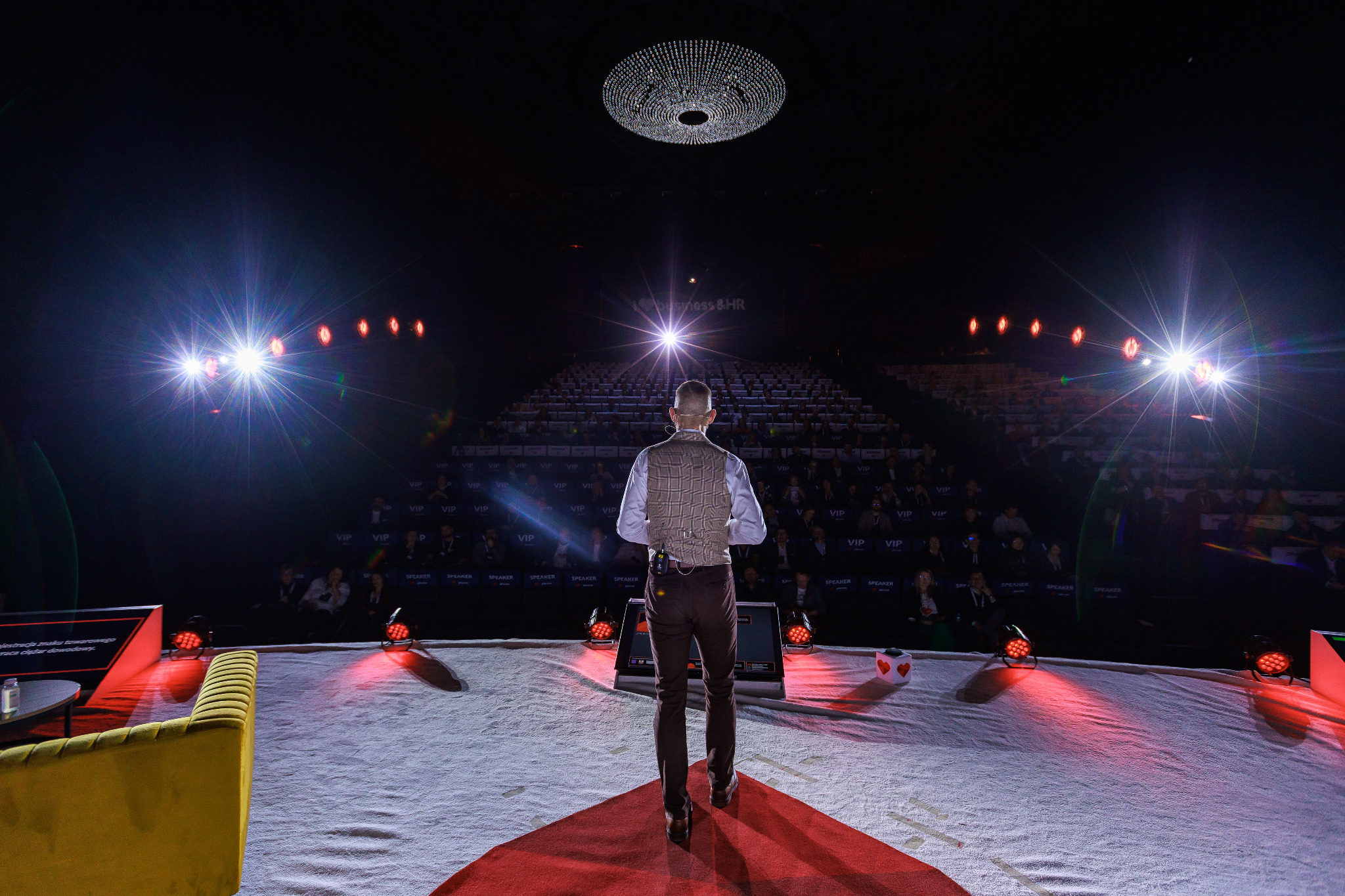Prelegent w brązowej kamizelce i spodniach, stojący tyłem na scenie oświetlonej reflektorami, z widownią w tle i żyrandolem nad głową, podczas konferencji biznesowej.