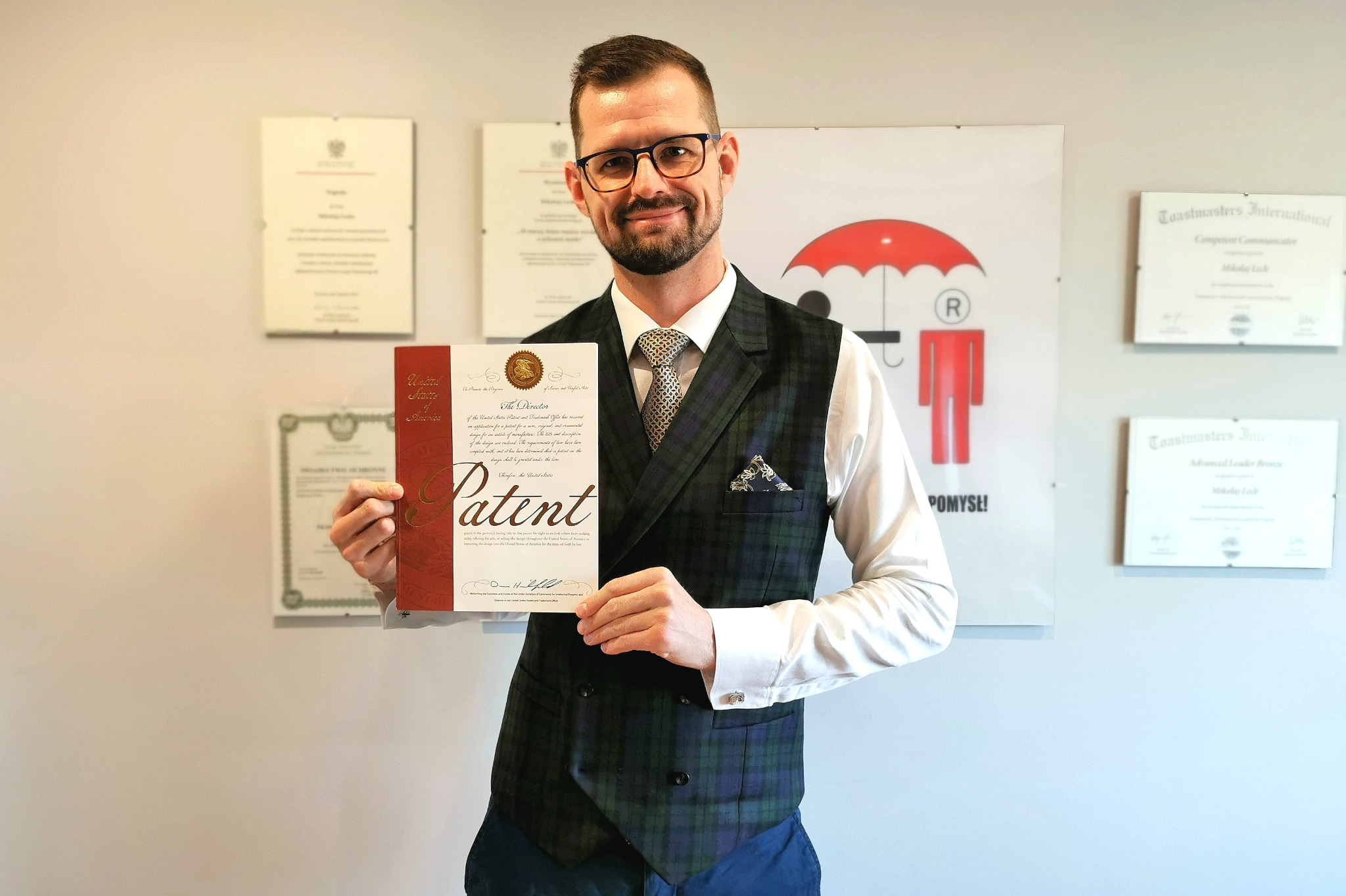 Mężczyzna w okularach i kamizelce prezentuje certyfikat patentowy na tle ściany z dyplomami i logo firmy.