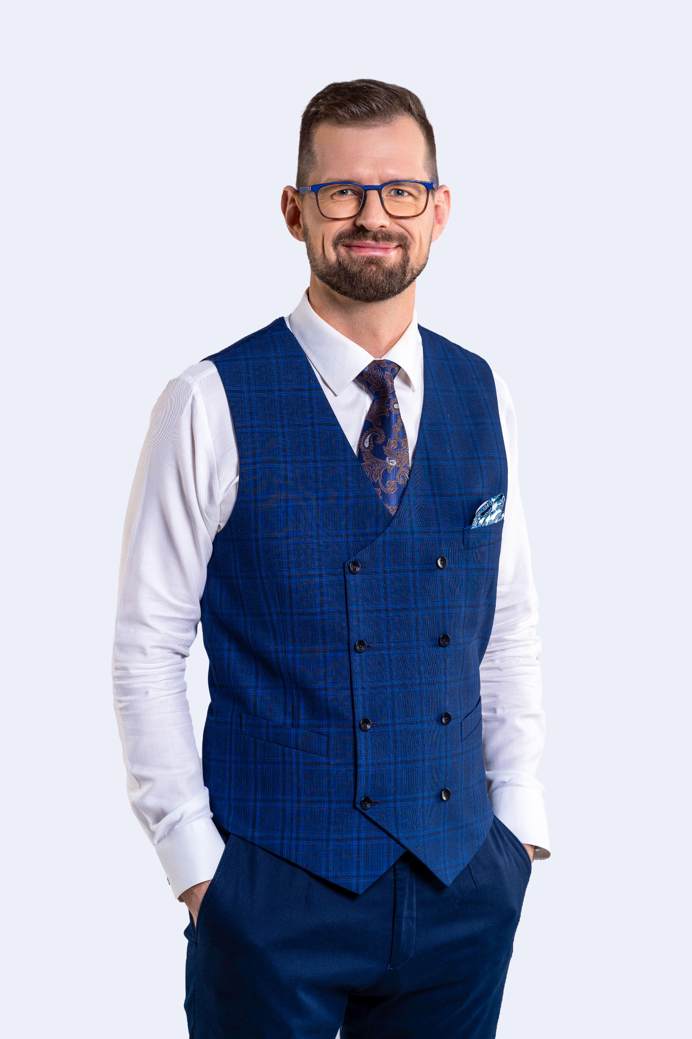 Uśmiechnięty prawnik w okularach i eleganckiej niebieskiej kamizelce w kratę, z chusteczką w kieszonce i wzorzystym krawatem, pozujący na jasnym tle.