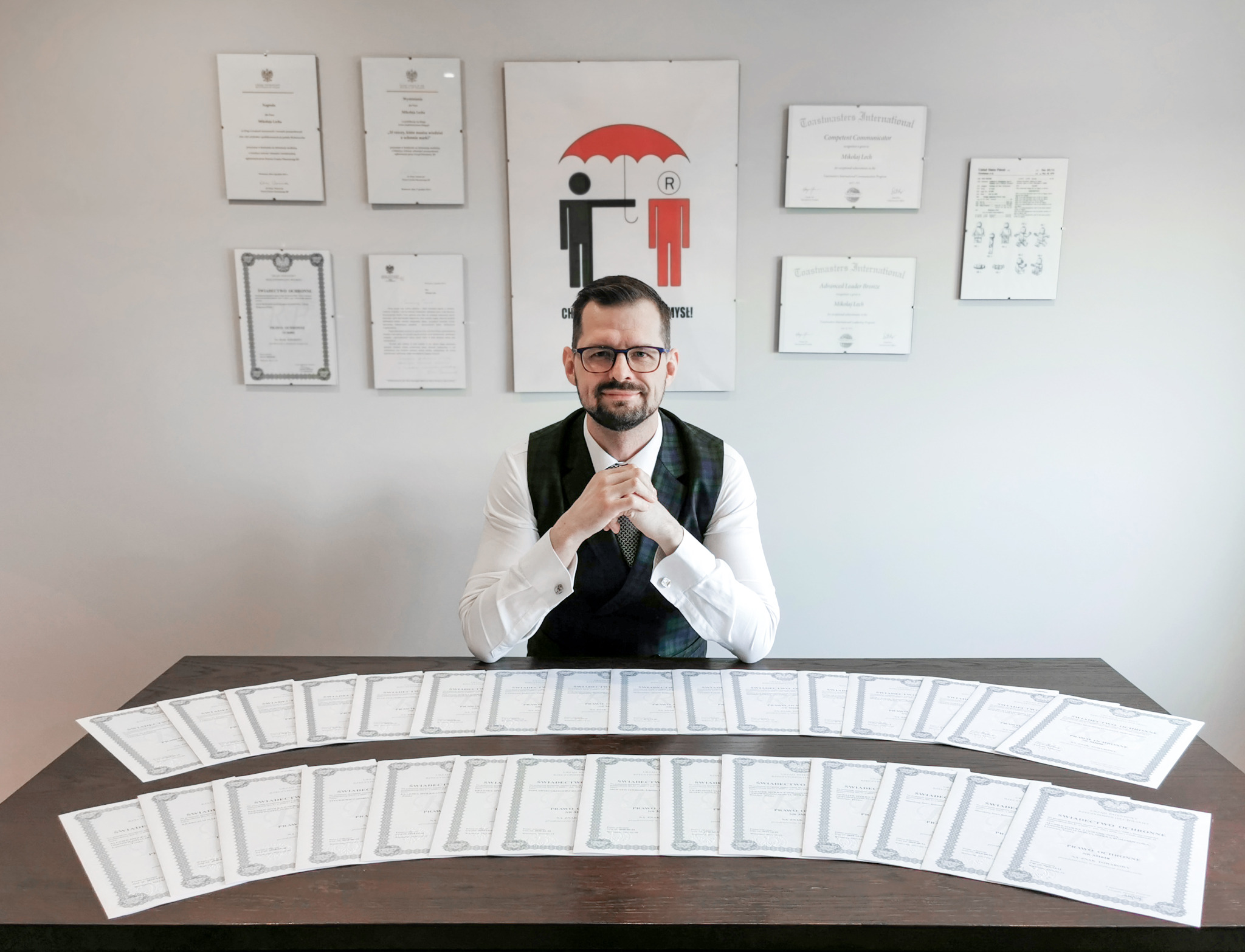 Portret prawnika w okularach za biurkiem z licznymi certyfikatami i dokumentami prawnymi, na ścianie dyplomy i logo firmy.