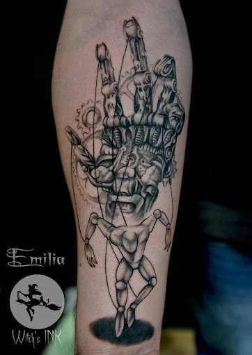 Tatuaż na przedramieniu przedstawiający mechaniczną dłoń marionetkarza kontrolującą kukiełkę, wykonany w Toruniu w stylu realistycznym z elementami steampunk.