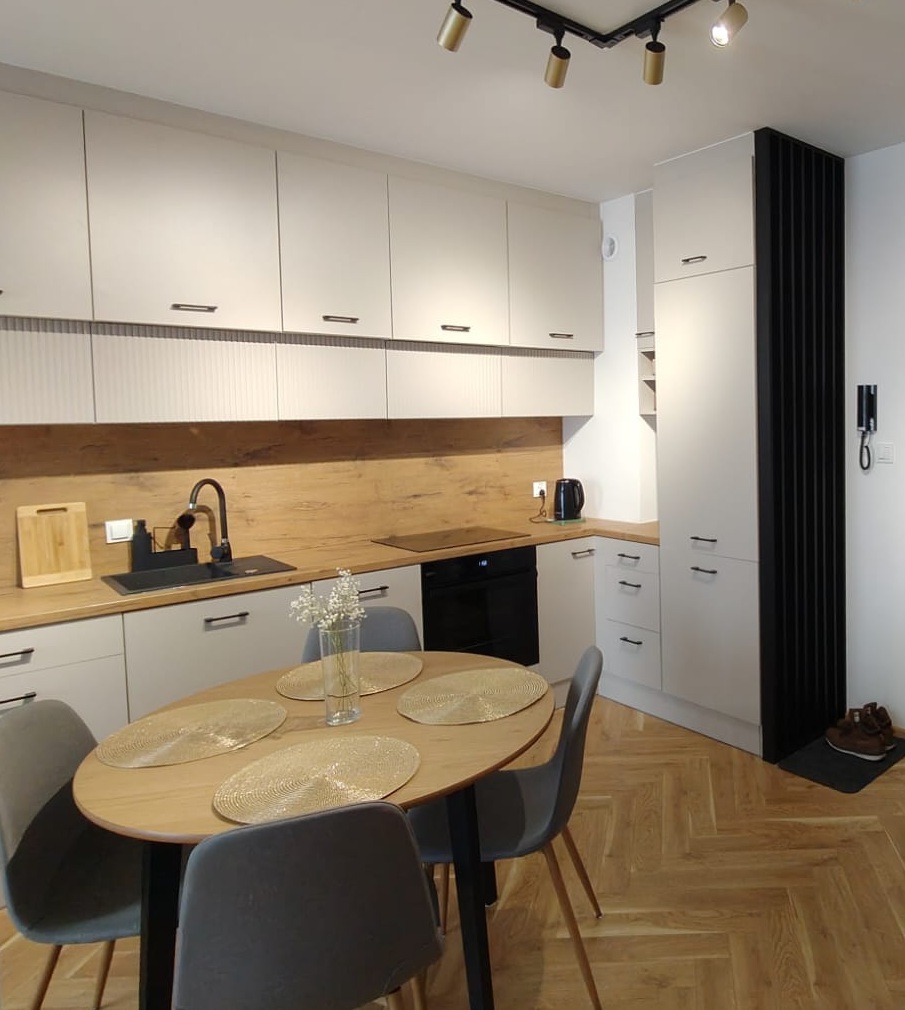 Jasna kuchnia z drewnianymi akcentami: okrągły stół z szarymi krzesłami, drewniany blat, białe szafki i parkiet w jodełkę. Minimalistyczny design z oświetleniem punktowym.