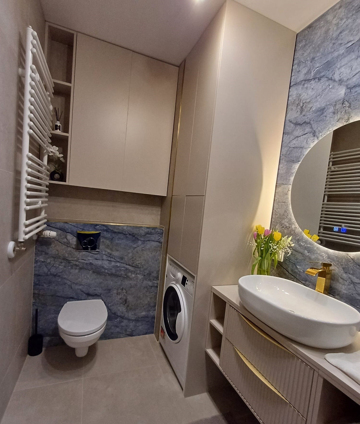 Nowoczesna łazienka z umywalką nablatową, marmurową ścianą i okrągłym lustrem z podświetleniem LED. Widoczny grzejnik drabinkowy i pralka.