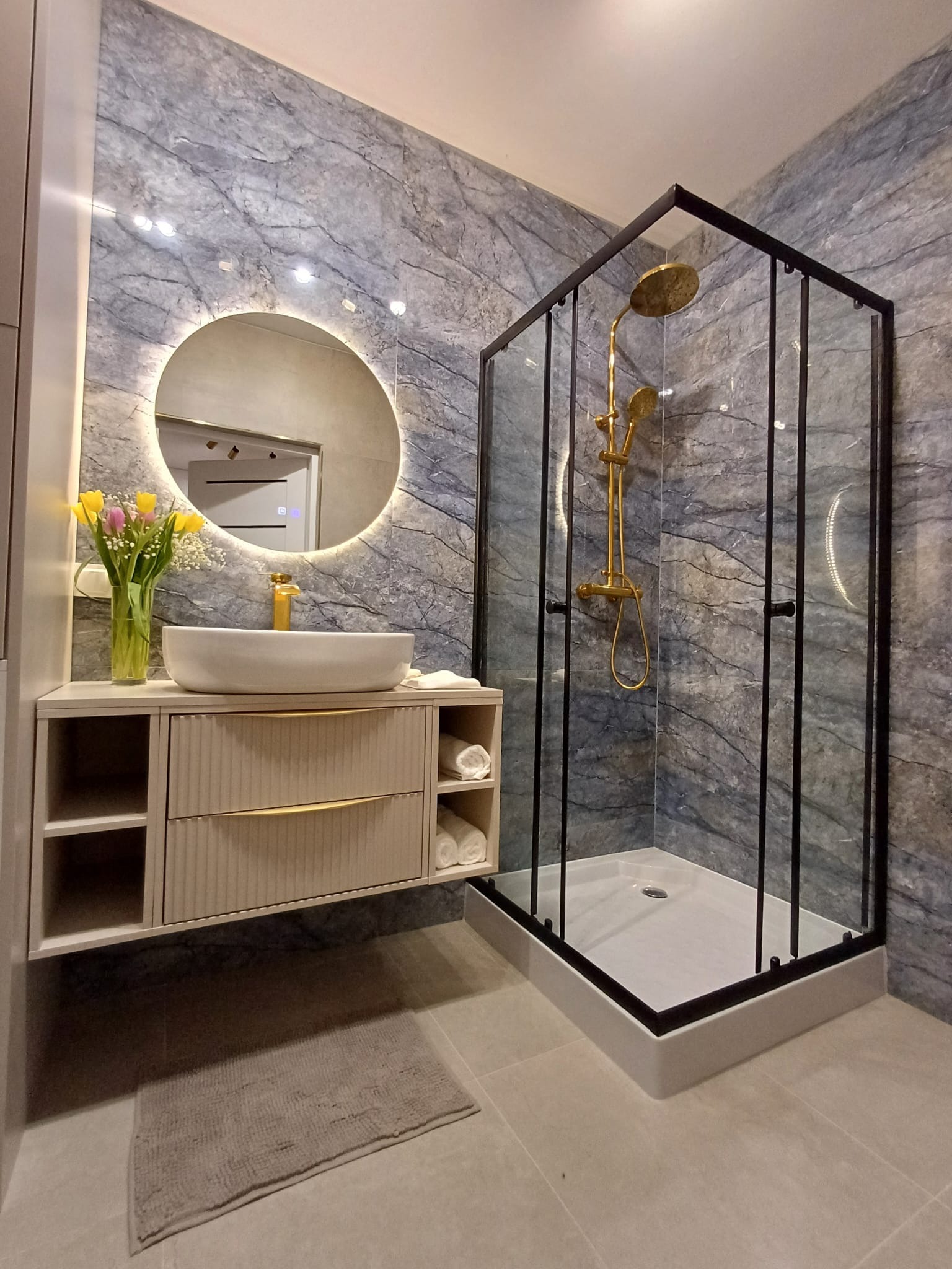 Nowoczesna łazienka z marmurową ścianą, okrągłym lustrem z podświetleniem LED, umywalką nablatową i kabiną prysznicową ze złotą armaturą. Minimalistyczny design.