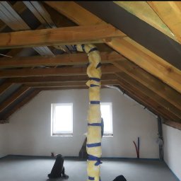 Renomo - Poddasze w trakcie adaptacji: widoczne drewniane krokwie, ocieplony pion wentylacyjny owinięty niebieską taśmą, w tle dwa okna, wylewka na podłodze zabezpieczona taśmą malarską.