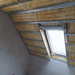 Renomo - Poddasze w trakcie izolacji: widoczna wełna mineralna pomiędzy metalowymi profilami, okno dachowe z białymi ramami, surowa ściana w tle.