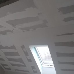 Renomo - Poddasze w trakcie prac wykończeniowych, widoczne fragmenty sufitu i ściany pokryte gładzią szpachlową, okno dachowe z widokiem na drzewa bez liści.