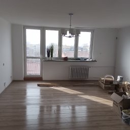 Renomo - Pusty pokój z nową, jasną podłogą panelową, z widocznymi prostopadłymi promieniami słonecznymi, białymi ścianami i oknami, z przygotowanymi materiałami i narzędziami do dalszych prac...