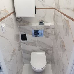 Renomo - Nowoczesna, wąska toaleta w wykończeniu marmurowym, z podtynkową spłuczką i minimalistycznym przyciskiem, oświetlona od góry, z widocznym fragmentem ramy drzwi.