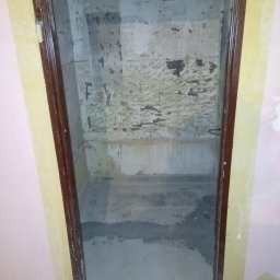 Renomo - Łazienka w trakcie demontażu starych płytek i przygotowania podłoża, widoczne ślady po kleju i resztki starej okładziny na ścianach, częściowo wylany podkład na podłodze, ościeżnica drzwiowa...