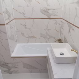 Renomo - Biała łazienka z marmurowymi płytkami, prostokątną umywalką nablatową i złotą armaturą, gdzie widoczny jest detal połączenia płytek z poziomym paskiem w kolorze miedzi.