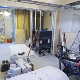 Renomo - Wnętrze w trakcie remontu, widoczne metalowe profile ścian działowych, materiały budowlane, narzędzia,  instalacje elektryczne pod sufitem i fragment starej ściany z cegły.