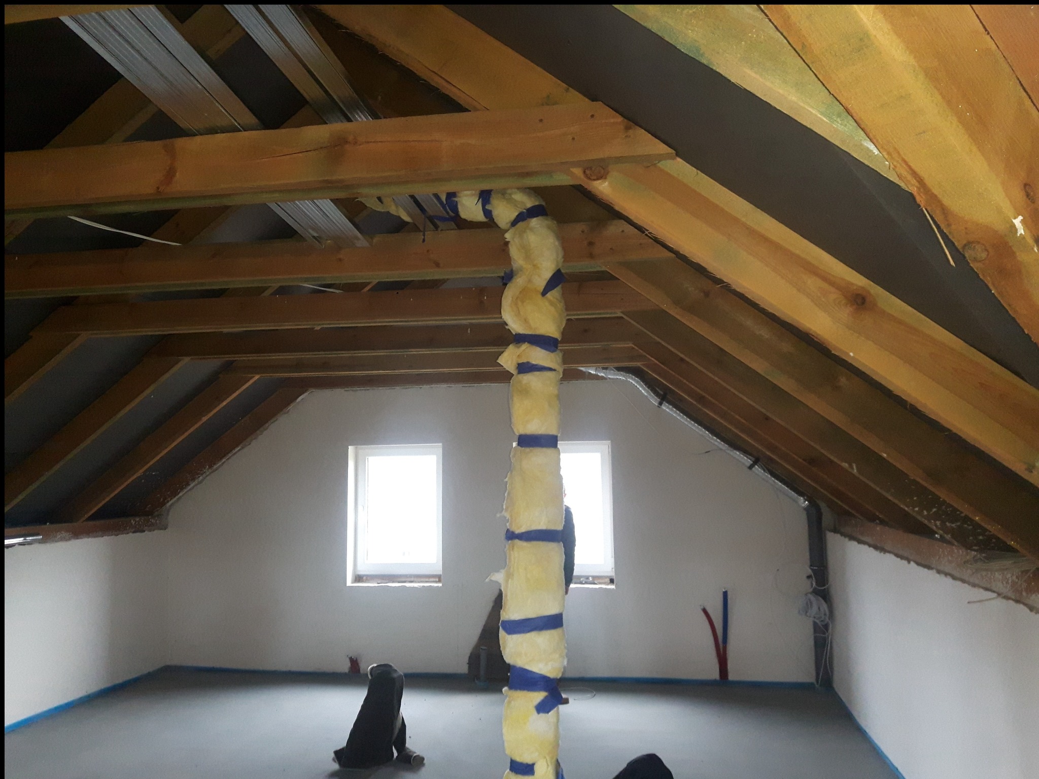 Poddasze w trakcie adaptacji: widoczne drewniane krokwie, ocieplony pion wentylacyjny owinięty niebieską taśmą, w tle dwa okna, wylewka na podłodze zabezpieczona taśmą malarską.