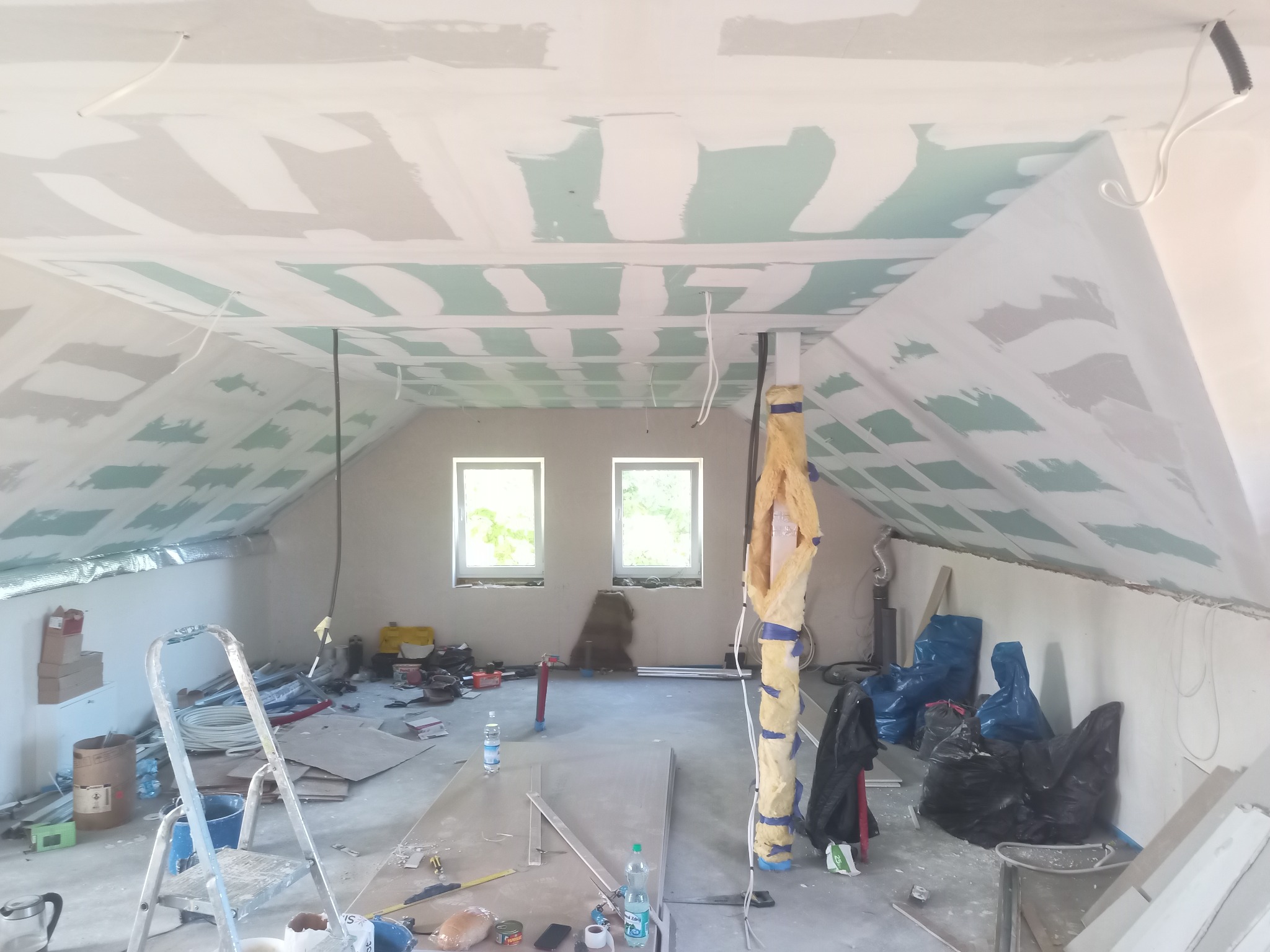 Poddasze w trakcie prac wykończeniowych: widoczne płyty gipsowo-kartonowe na skosach sufitu, częściowo zaszpachlowane, instalacja elektryczna, ocieplony filar, okna dachowe i bałagan budowlany...