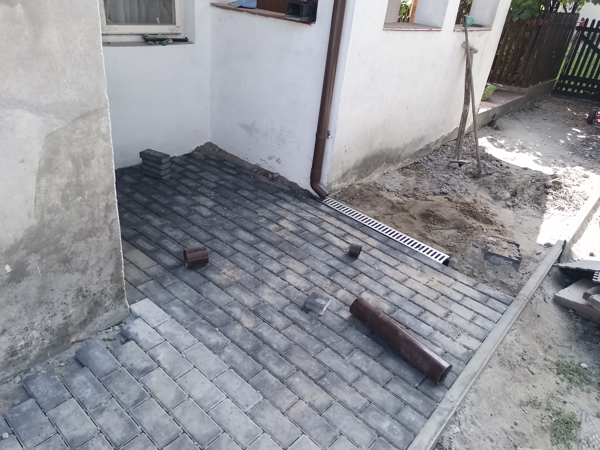 Układanie szarej kostki brukowej na tarasie zewnętrznym przy budynku, z widocznym odpływem liniowym i fragmentami niezakończonej nawierzchni.