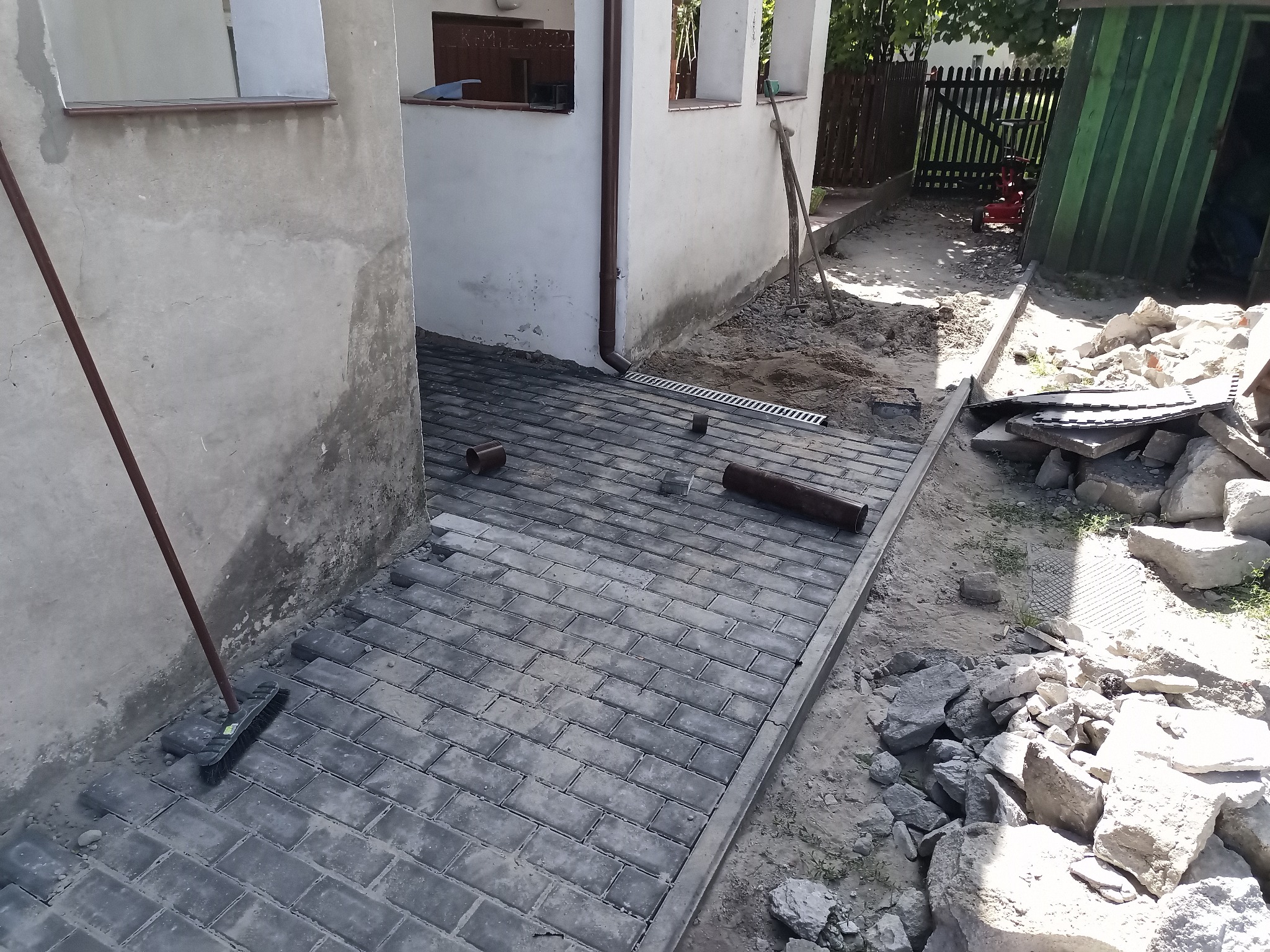 Układanie szarej kostki brukowej na posesji, widoczne niedokończone prace, krawężniki, przygotowane podłoże i gruz obok.