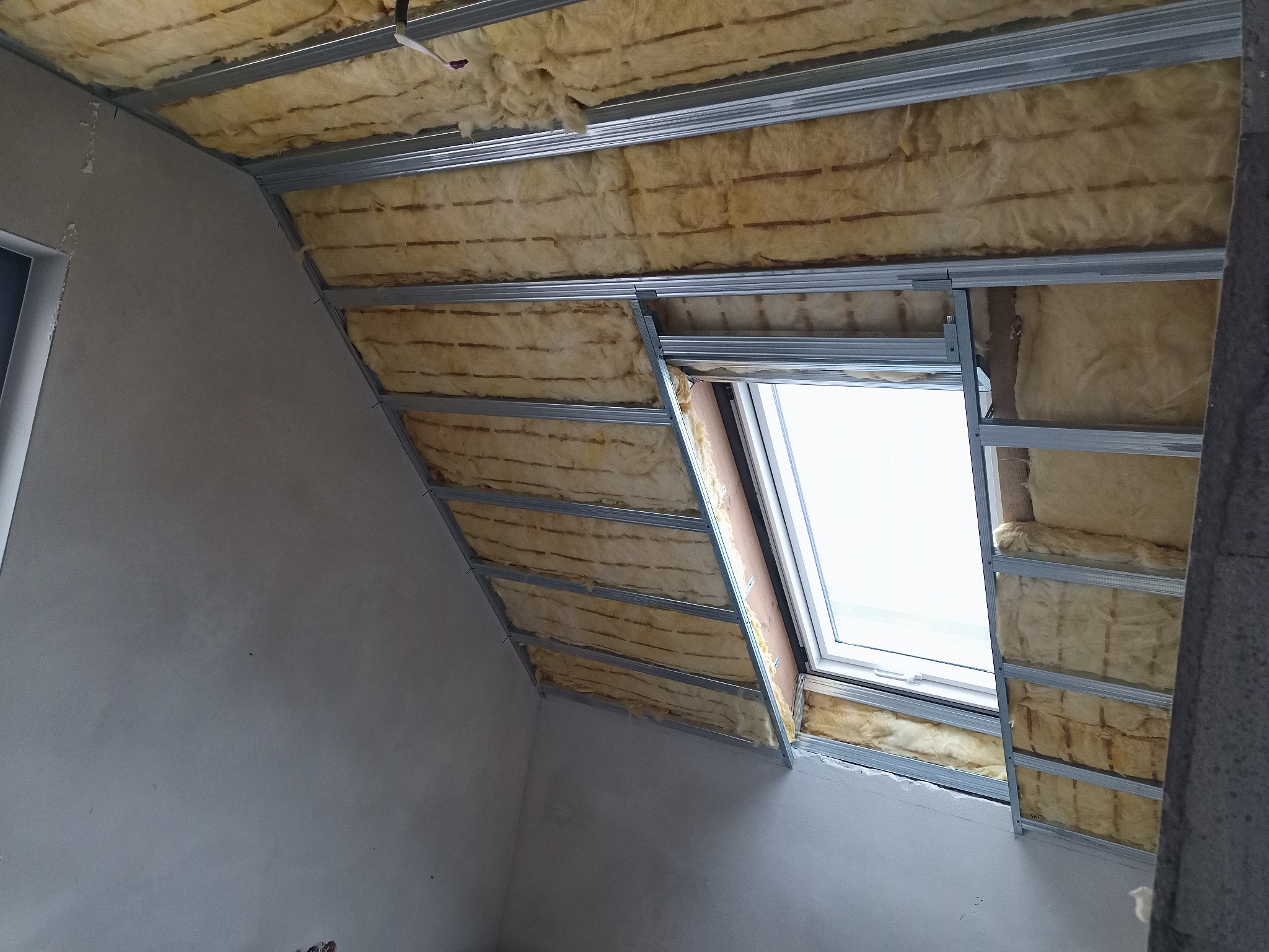 Poddasze w trakcie izolacji: widoczna wełna mineralna pomiędzy metalowymi profilami, okno dachowe z białymi ramami, surowa ściana w tle.