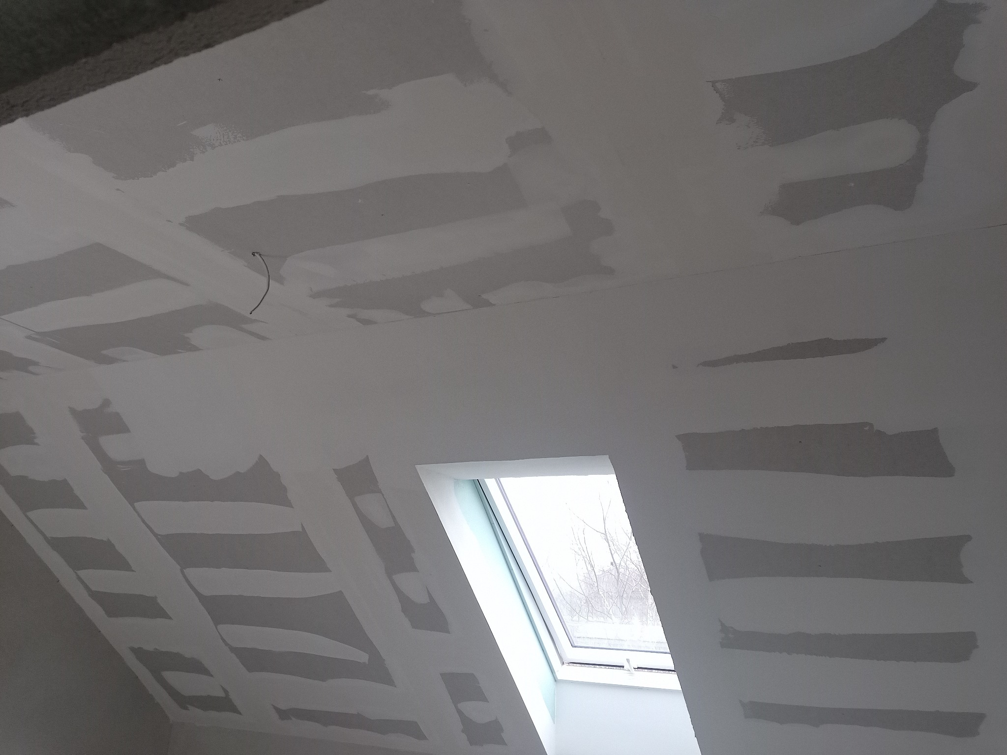 Poddasze w trakcie prac wykończeniowych, widoczne fragmenty sufitu i ściany pokryte gładzią szpachlową, okno dachowe z widokiem na drzewa bez liści.