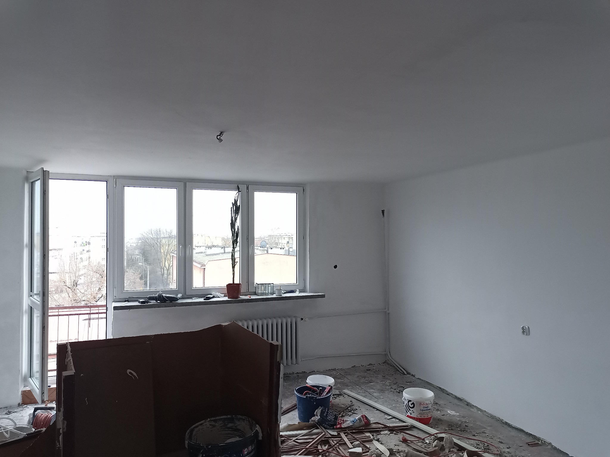 Pokój w trakcie prac wykończeniowych: świeżo pomalowane ściany na biało, widoczne puszki z farbą, resztki materiałów budowlanych na podłodze, okno z rośliną doniczkową, zabezpieczone kartonem...