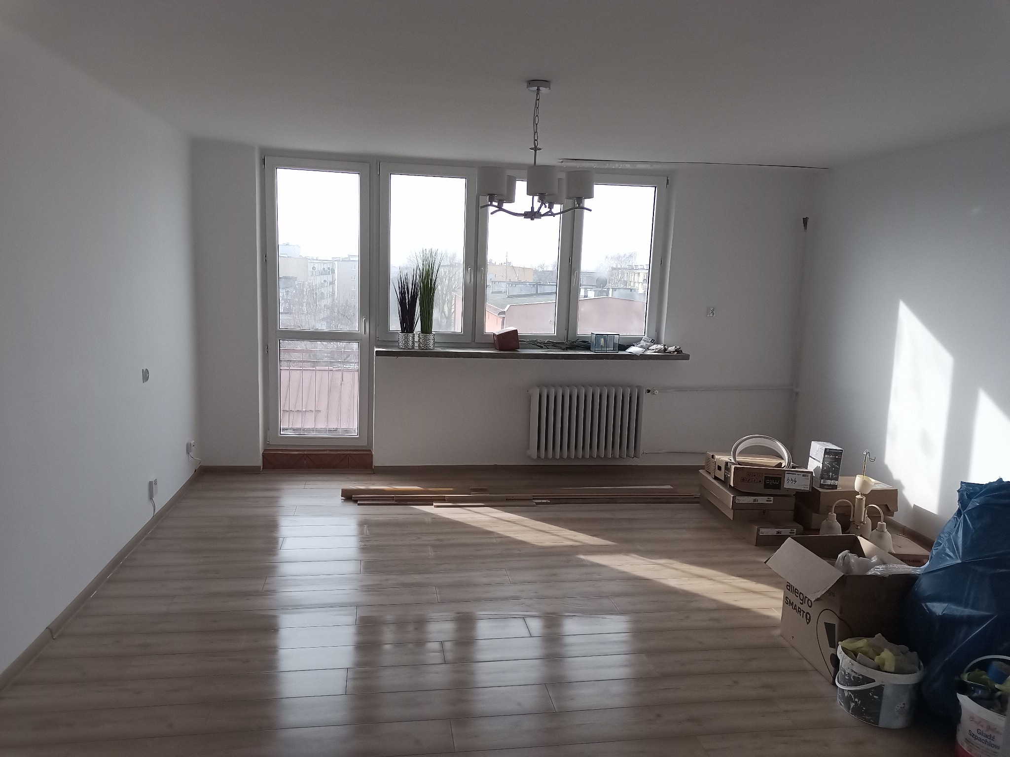 Pusty pokój z nową, jasną podłogą panelową, z widocznymi prostopadłymi promieniami słonecznymi, białymi ścianami i oknami, z przygotowanymi materiałami i narzędziami do dalszych prac...