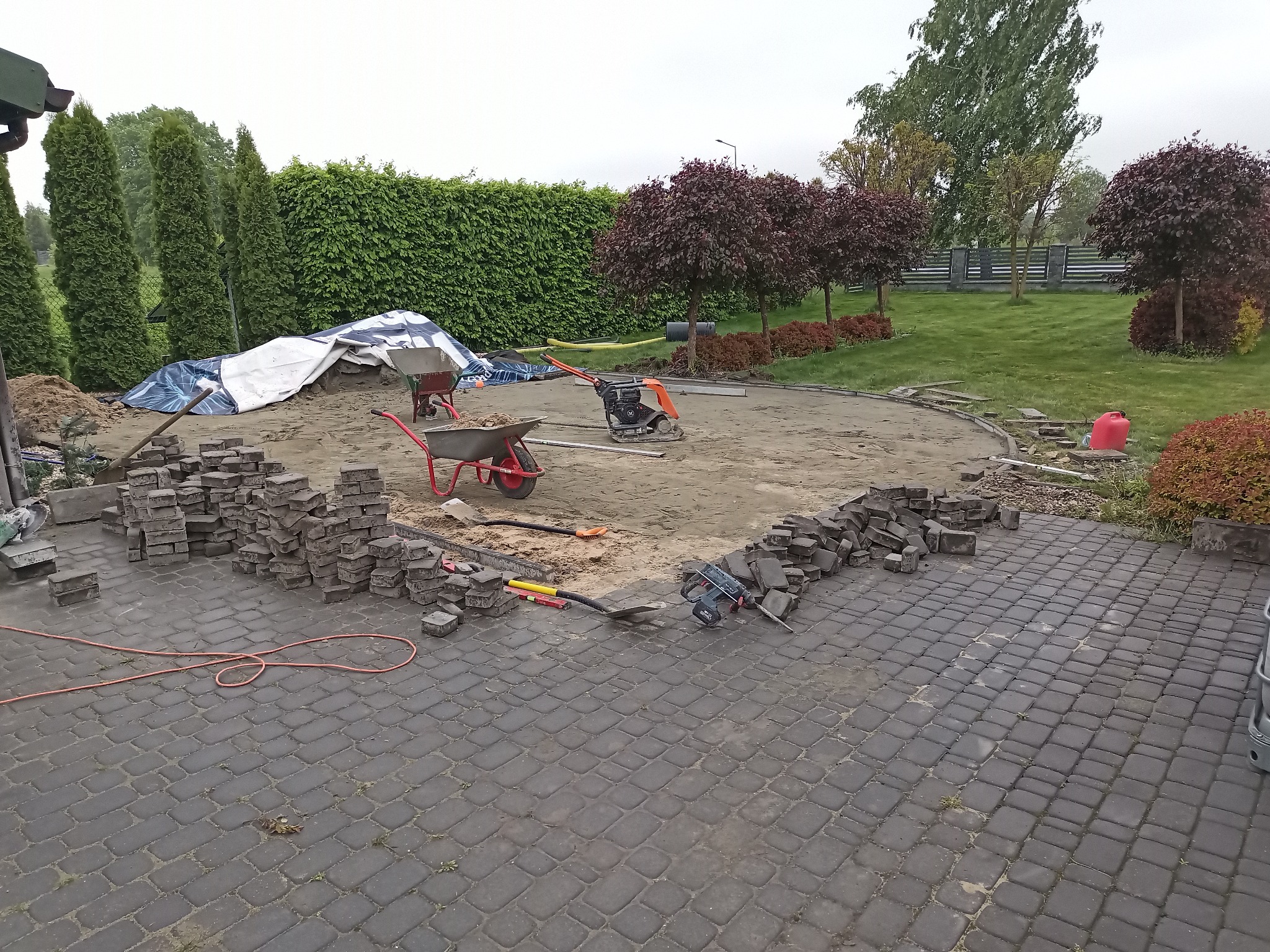 Układanie kostki brukowej na posesji: widoczne narzędzia, kostka, ubijarka, dwie taczki i teren przygotowany pod dalsze prace.