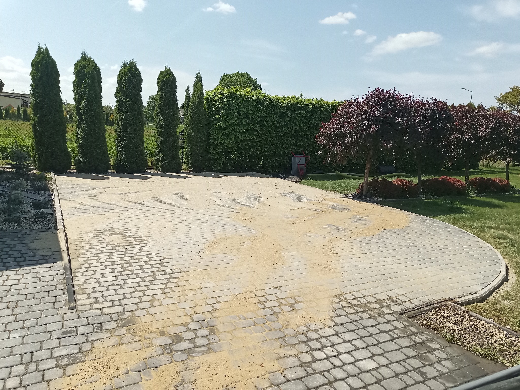 Ułożona kostka brukowa na podjeździe, częściowo zasypana piaskiem, z widocznymi drzewami i krzewami w tle.