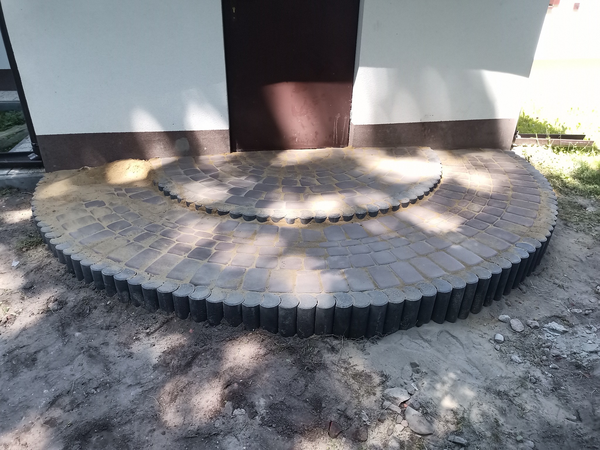 Półokrągłe schody wejściowe wyłożone szarą kostką brukową z obrzeżem z betonowych palisad, piasek między kostkami, brązowe drzwi wejściowe w tle.