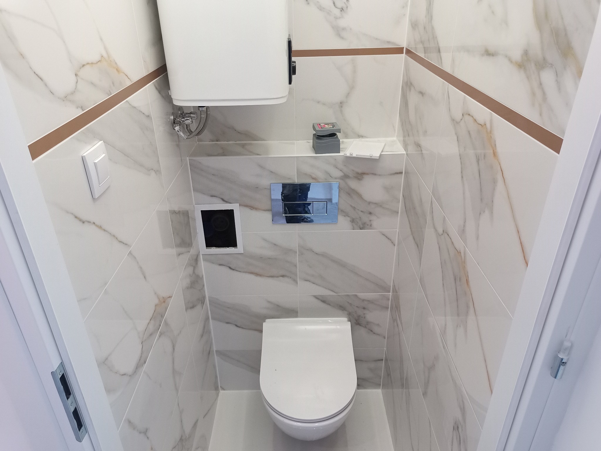 Nowoczesna, wąska toaleta w wykończeniu marmurowym, z podtynkową spłuczką i minimalistycznym przyciskiem, oświetlona od góry, z widocznym fragmentem ramy drzwi.