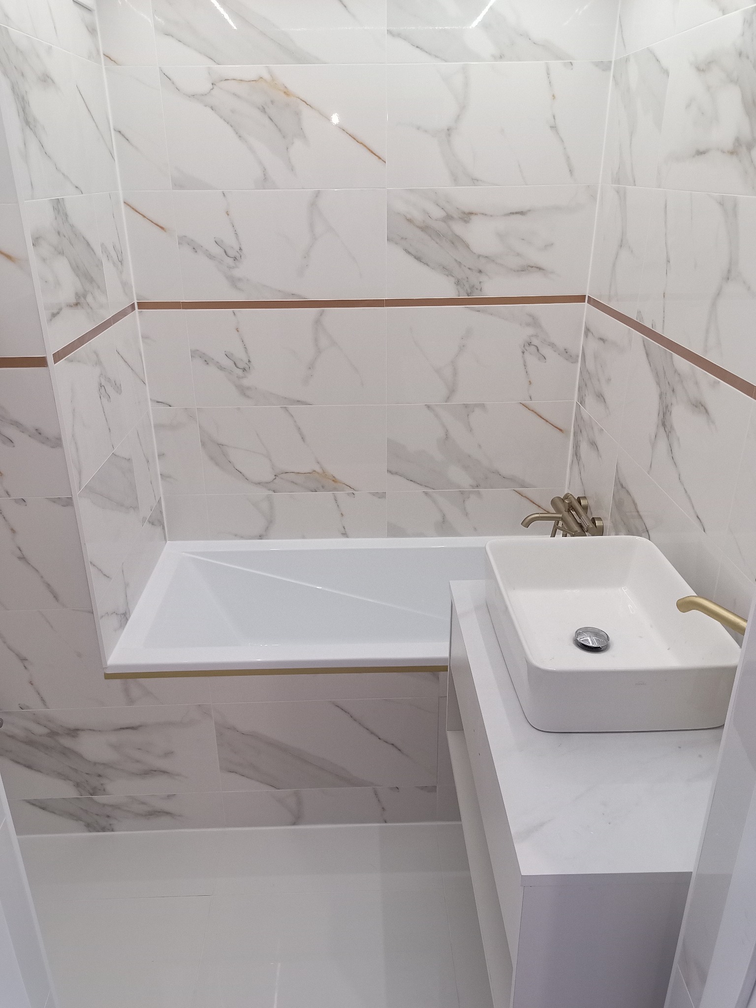 Biała łazienka z marmurowymi płytkami, prostokątną umywalką nablatową i złotą armaturą, gdzie widoczny jest detal połączenia płytek z poziomym paskiem w kolorze miedzi.