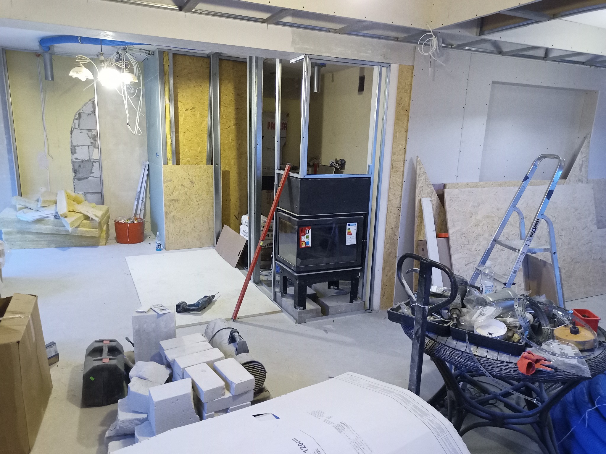 Wnętrze w trakcie remontu, widoczne metalowe profile ścian działowych, materiały budowlane, narzędzia,  instalacje elektryczne pod sufitem i fragment starej ściany z cegły.