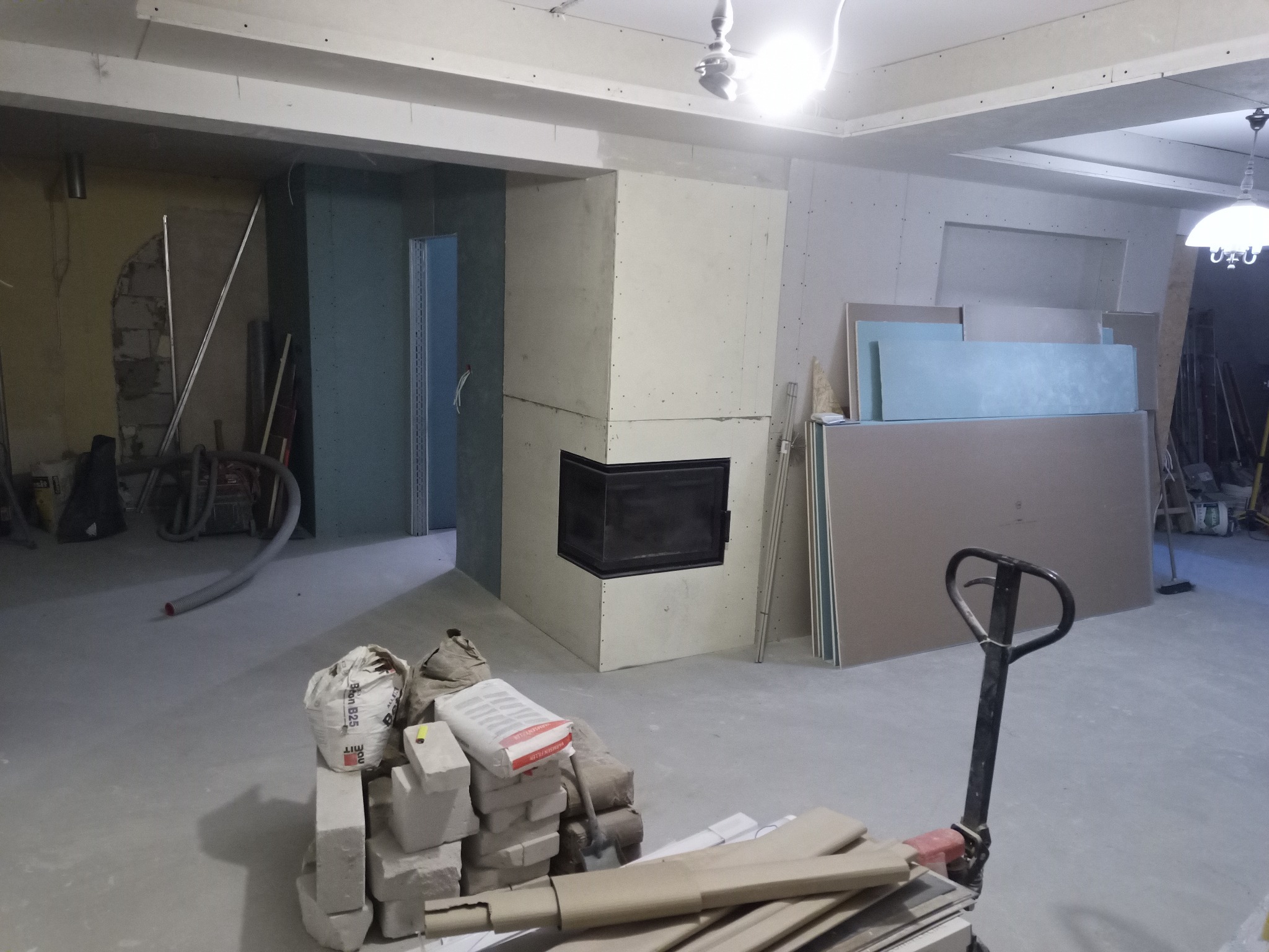 Wnętrze w trakcie prac wykończeniowych: zabudowa kominka z płyt gipsowo-kartonowych, worki z zaprawą murarską i paleta z płytami na podłodze, widoczne ściany przygotowane pod gładzie.