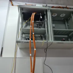 Szafa serwerowa z zamontowanym sprzętem sieciowym, widoczne pomarańczowe kable uporządkowane taśmą, wystające kable zasilające, surowy betonowy sufit w tle.