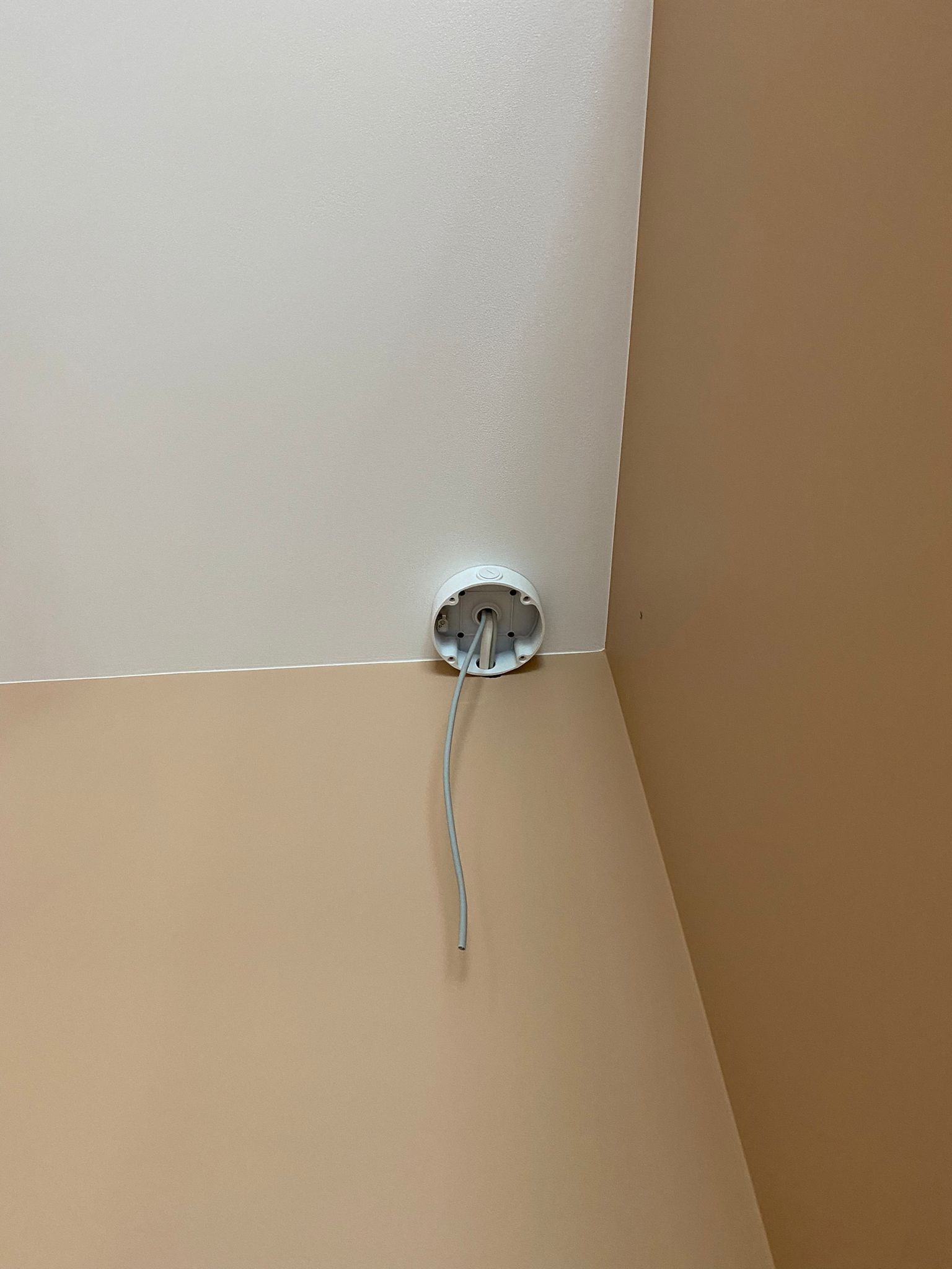 Biała puszka montażowa na suficie, z wystającymi kablami, widok z dołu w rogu pomieszczenia o dwóch odcieniach beżu.