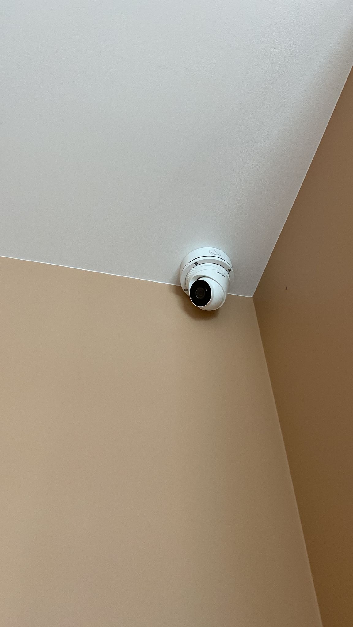 Kopułkowa kamera monitoringu Hikvision zamontowana na białym suficie w rogu pomieszczenia o beżowych ścianach.