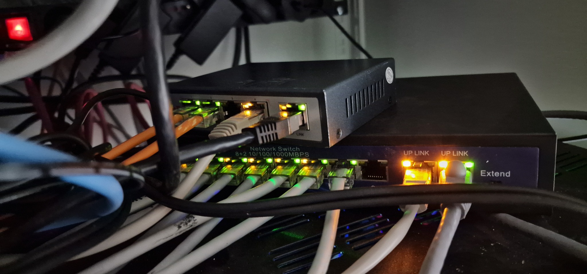 Dwa przełączniki sieciowe z podłączonymi kablami Ethernet, diody LED sygnalizują aktywność połączeń, widoczne oznaczenia portów 'UP LINK' i specyfikacja '8+2 10/100/1000MBPS'.