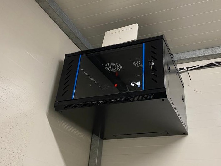 Czarna szafa rackowa z niebieskimi akcentami, zamontowana na białej ścianie, z routerem na górze i widocznymi kablami. Kluczyk w zamku szafy.