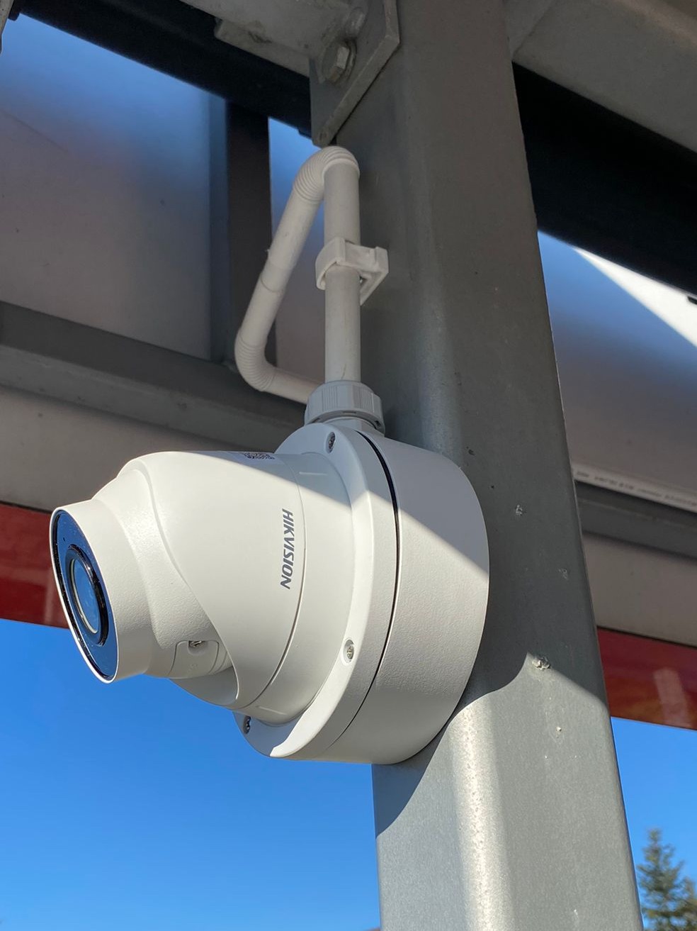 Kamera Hikvision typu dome, zamontowana na szarej, metalowej konstrukcji z widocznymi przewodami w białej rurce PCV, na tle błękitnego nieba.