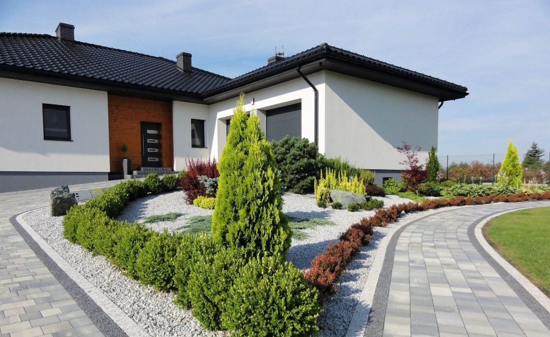 Nowoczesny ogród z krzewami, drzewkami iglastymi i ścieżką z kostki brukowej wokół domu z białymi ścianami i ciemnym dachem.