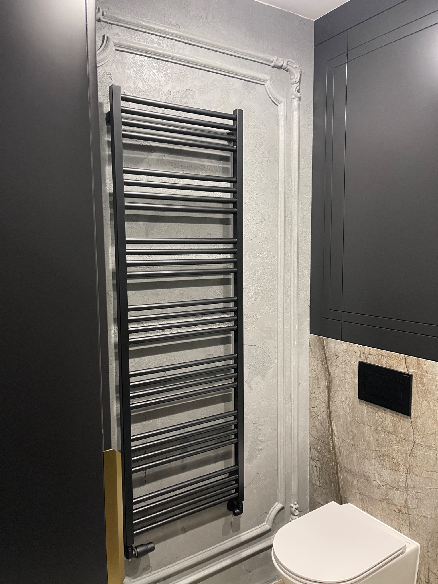 Nowoczesna łazienka z czarnym grzejnikiem drabinkowym na ścianie z dekoracyjnym stiukiem, obok zabudowa meblowa w kolorze grafitowym i toaleta z deską wolnoopadającą.