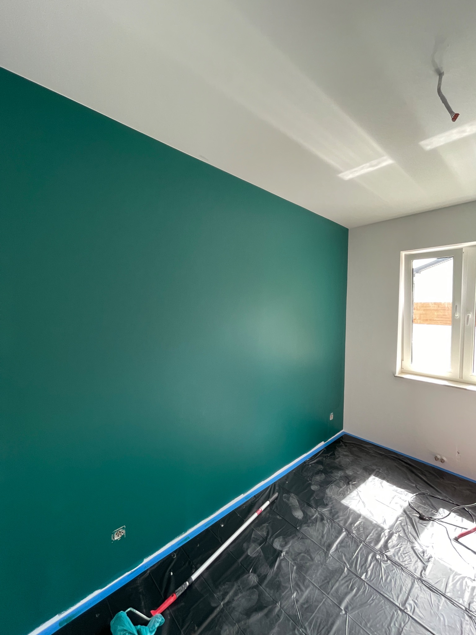 Świeżo pomalowana ściana w odcieniu butelkowej zieleni, z widoczną taśmą malarską przy podłodze i zabezpieczoną folią podłogą, w rogu pokoju widoczne okno.