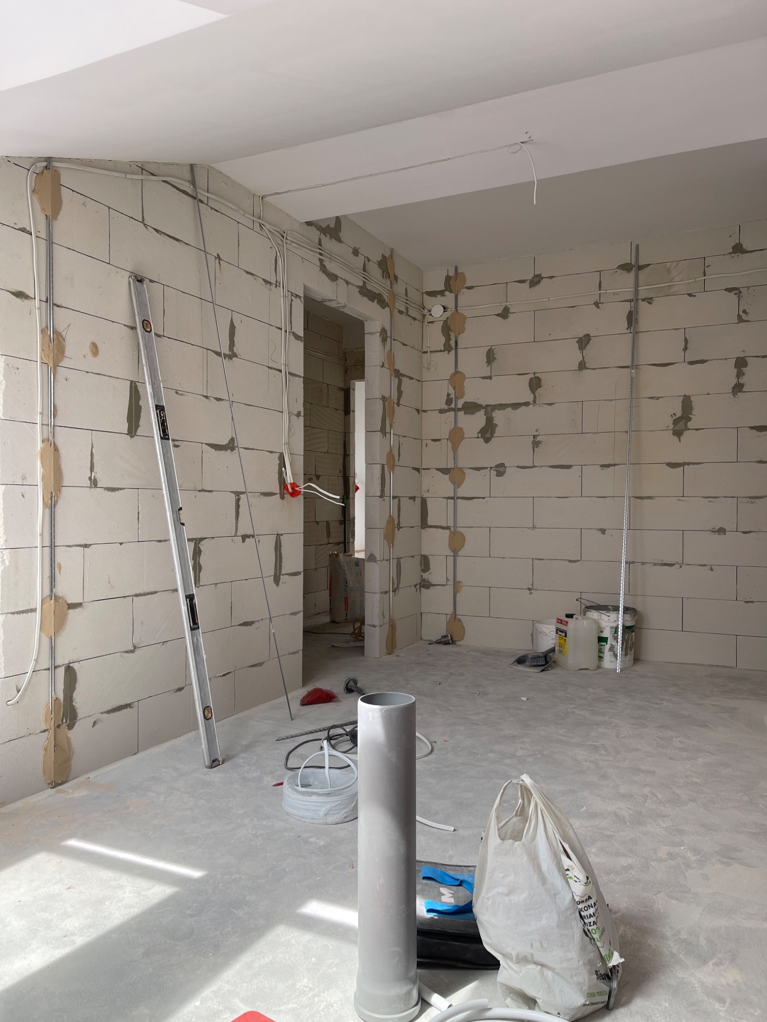 Surowy stan wnętrza w trakcie prac: widoczne kable elektryczne przymocowane do ściany z betonu komórkowego, poziomica oparta o ścianę, narzędzia i materiały budowlane na podłodze, sufit...