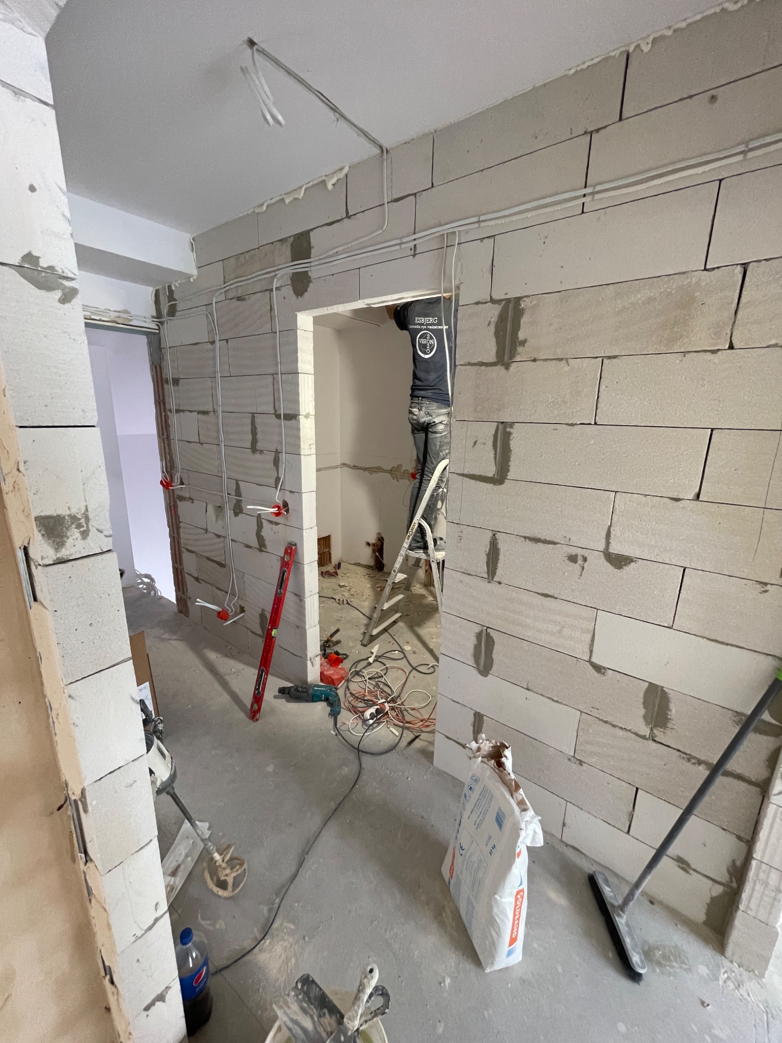 Wnętrze w trakcie budowy ściany z betonu komórkowego, widoczne narzędzia, poziomica, mieszadło do zaprawy oraz pracownik na drabinie w przejściu do innego pomieszczenia.
