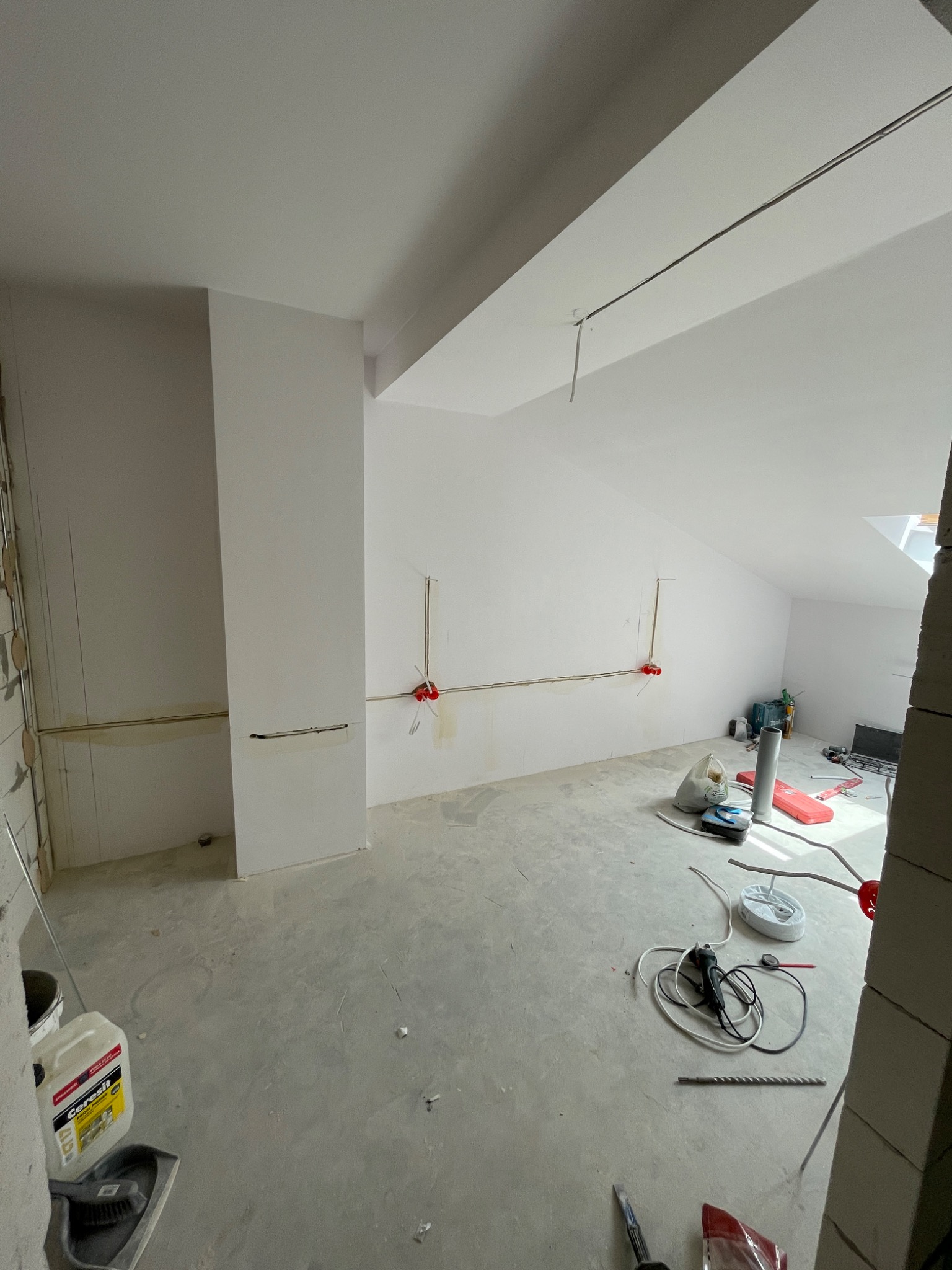 Wnętrze poddasza w trakcie prac wykończeniowych: widoczne przygotowane ściany pod gładzie, instalacja elektryczna, narzędzia i materiały budowlane na szarym, betonowym podłożu.