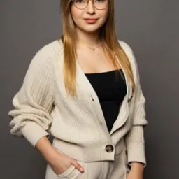 Portret kobiety w okularach, beżowym kardiganie i spodniach na szarym tle. Uśmiechnięta, z ręką w kieszeni. Stonowana kolorystyka, naturalne światło.