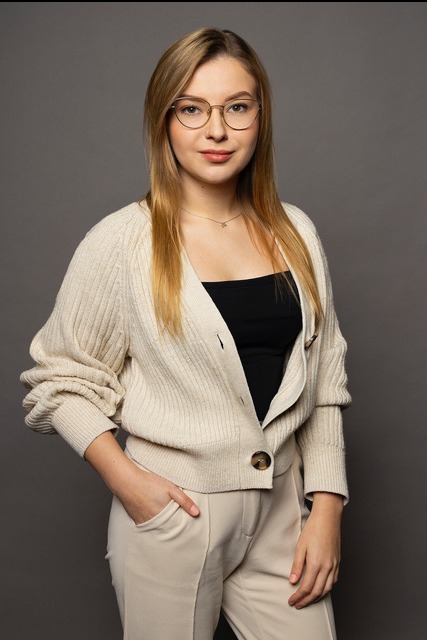 Portret kobiety w okularach, beżowym kardiganie i spodniach na szarym tle. Uśmiechnięta, z ręką w kieszeni. Stonowana kolorystyka, naturalne światło.