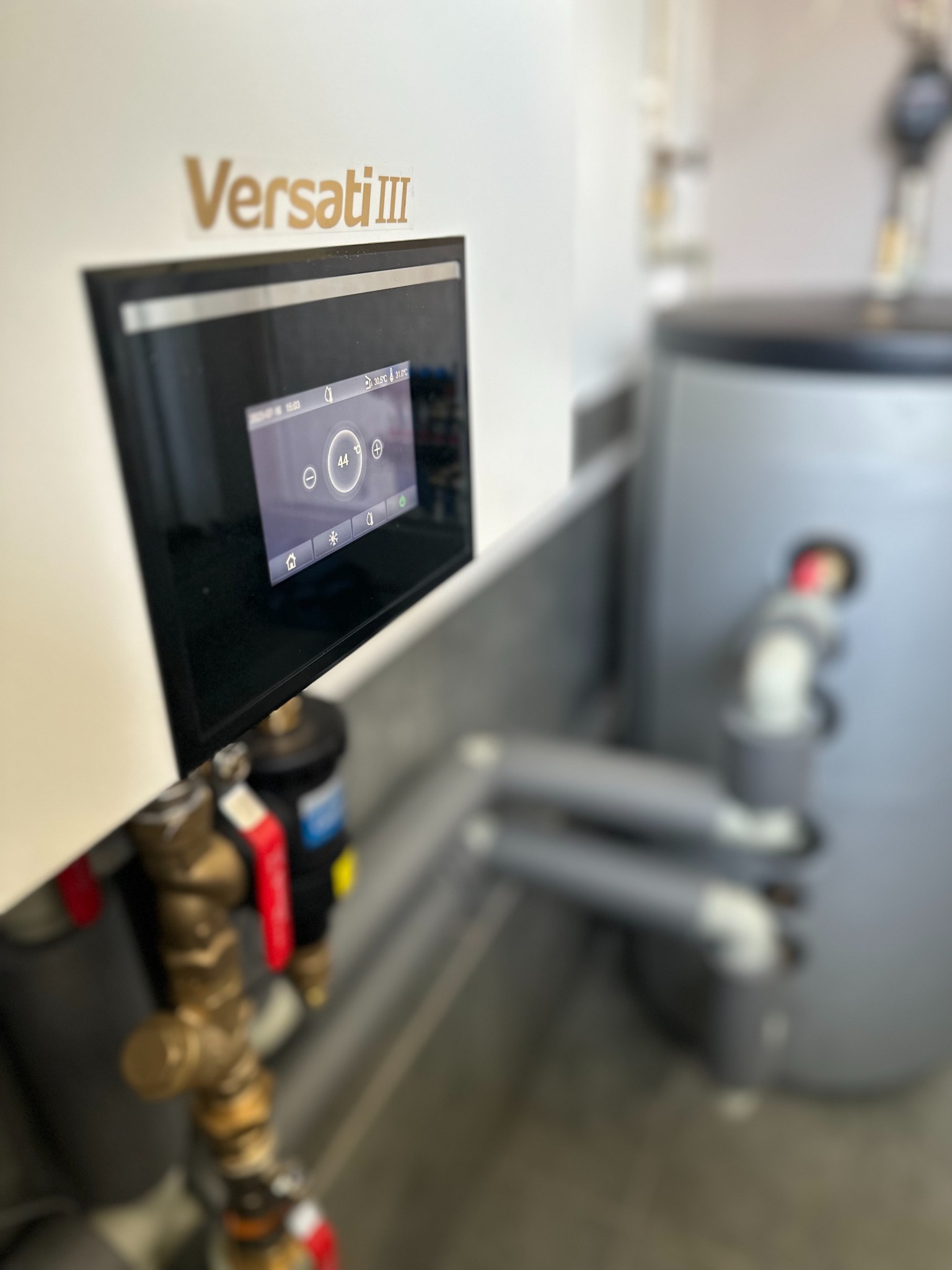 Pompa ciepła Versati III z widocznym panelem sterowania i podłączeniem hydraulicznym, zbiornik buforowy w tle.