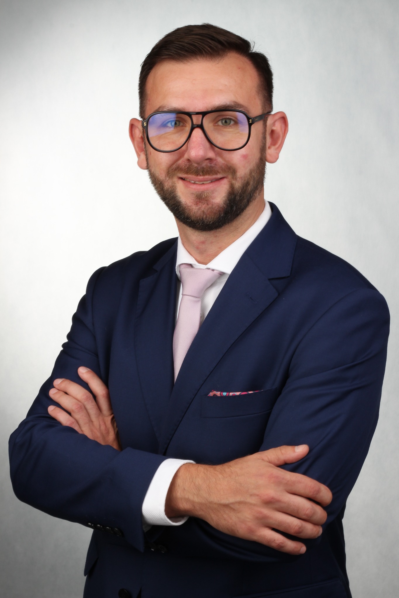 Portret uśmiechniętego mężczyzny w okularach i granatowym garniturze z różowym krawatem i poszetką w kieszonce, stojącego na jasnym tle z założonymi rękami.